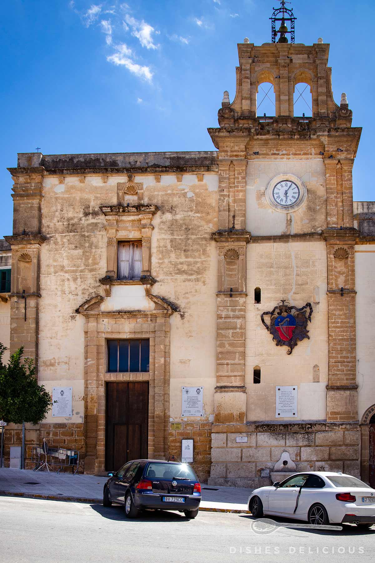 Turm mit Uhr und Wappen an einem historischen Gebäude unter blauem Himmel, davor zwei geparkte Autos.