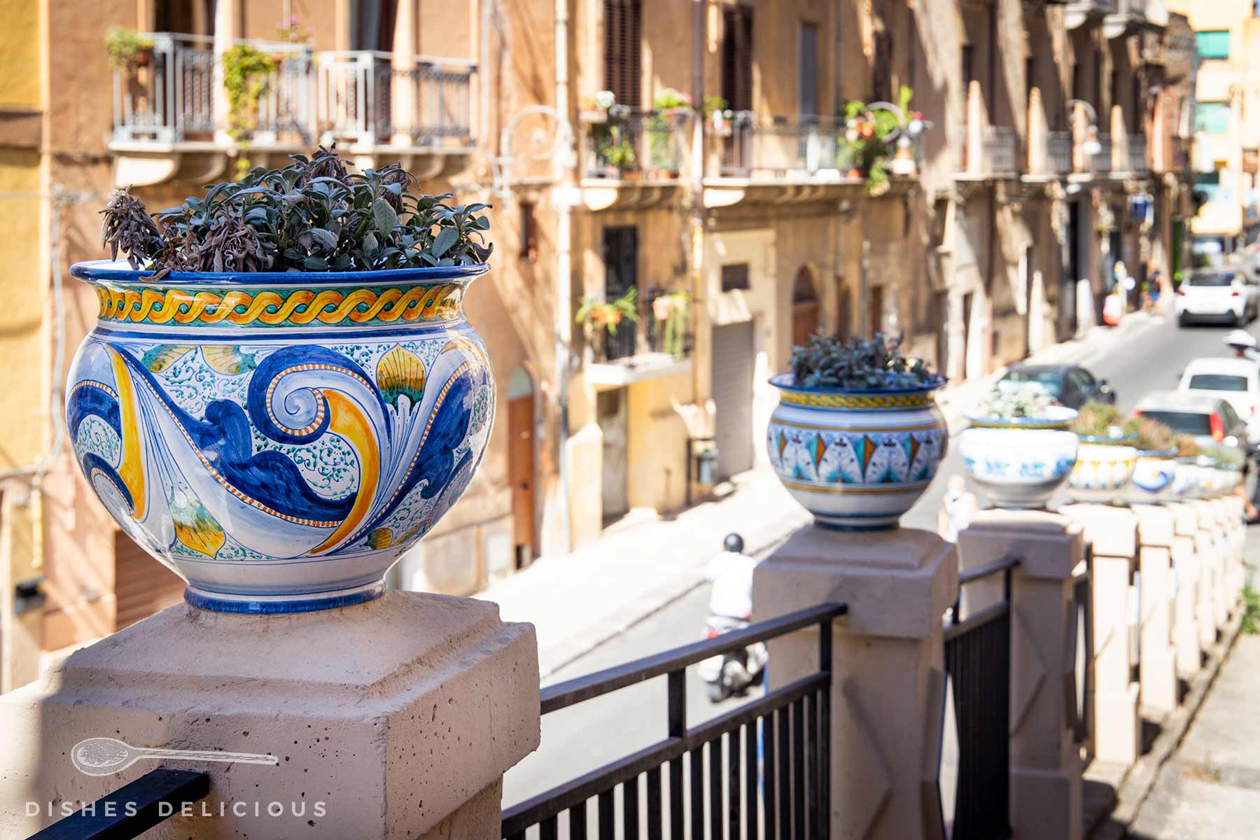 Mehrere kunstvoll bemalte Keramikvasen mit Pflanzen auf einer Steinmauer entlang einer sonnigen Straße in Sciacca.