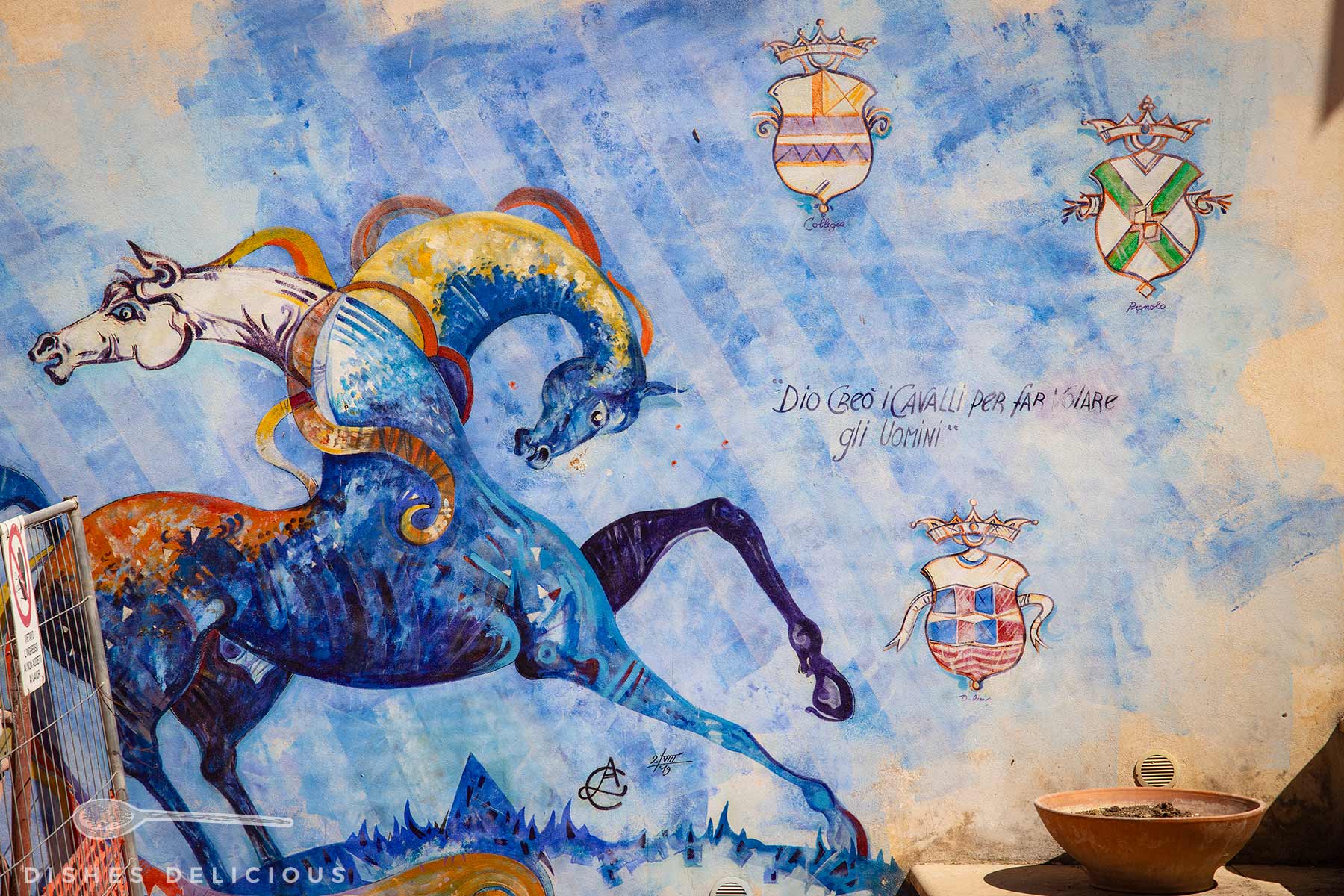Wandbemalung mit zwei Pferden, die die Köpfe in unterschiedliche Richtungen drehen. Drei Wappenschilder und Schriftzug 'Dio creò i cavalli per far volare gli uomini'.