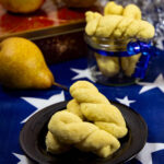 Drei gedrehte Affogagatti-Kekse auf einem schwarzen Teller, im Hintergrund weitere Kekse in einem Glasschälchen, daneben eine Birne auf einem blau-weißen Sternentuch.