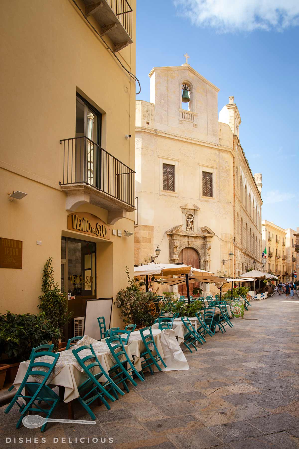 Straßencafé mit türkisfarbenen Stühlen vor einer historischen Kirche unter blauem Himmel.
