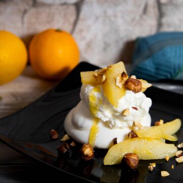 Baiser mit Sahne, Birnenscheiben und gehackten Haselnüssen auf schwarzem Teller, im Hintergrund zwei Orangen und ein blaues Tuch.