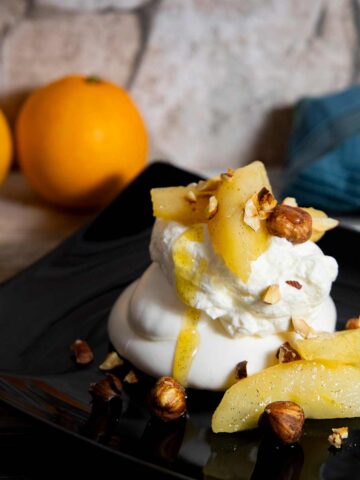 Weihnachtsdessert: Baiser mit karamellisierten Birnen und gerösteten Haselnüssen Baiser mit Sahne, Birnenscheiben und gehackten Haselnüssen auf schwarzem Teller, im Hintergrund zwei Orangen und ein blaues Tuch.