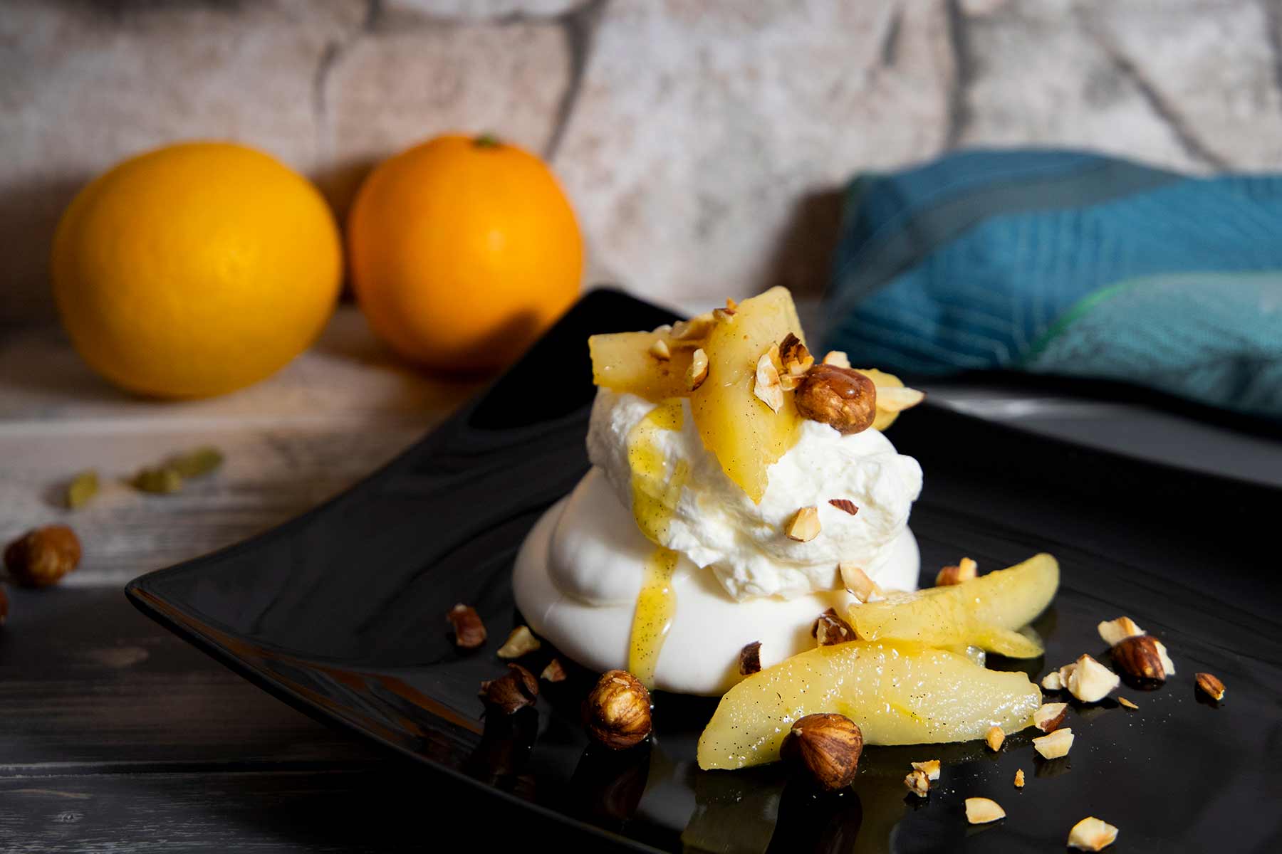 Baiser mit Sahne, Birnenscheiben und gehackten Haselnüssen auf schwarzem Teller, im Hintergrund zwei Orangen und ein blaues Tuch.