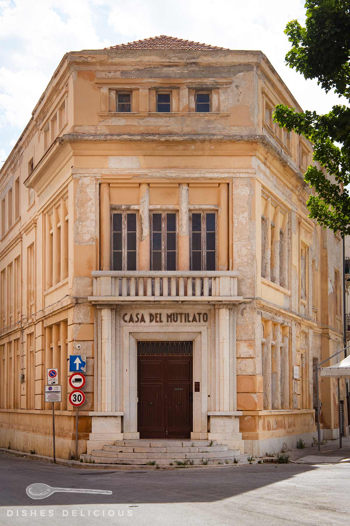 Leicht verwittertes Eckgebäude mit der Aufschrift 'Casa del Mutilato' über dem Eingang und Verkehrsschildern an der Straßenecke.