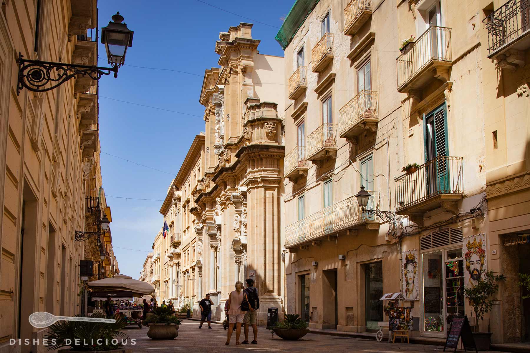 Sonniger Corso Vittorio Emanuele in Trapani mit barockem Gebäude, mehreren Balkonen und Passanten auf dem Pflasterweg.