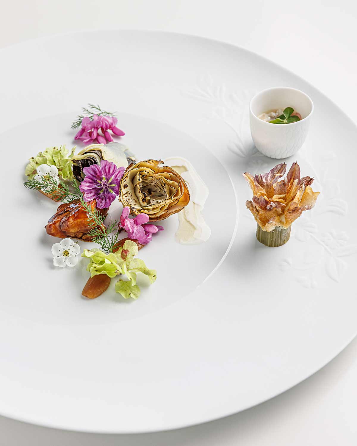 Auf weißem Teller angerichtete Speise mit Artischockenherzen, essbaren Blüten und einem kleinen weißen Becher mit Sauce.