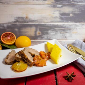 Vier Scheiben Entenbrust mit Blutorangenscheiben und zwei Polentastücken auf einem weißem Teller, daneben goldene Besteckgarnitur und ganze Zitrusfrüchte auf rotem Holztisch.