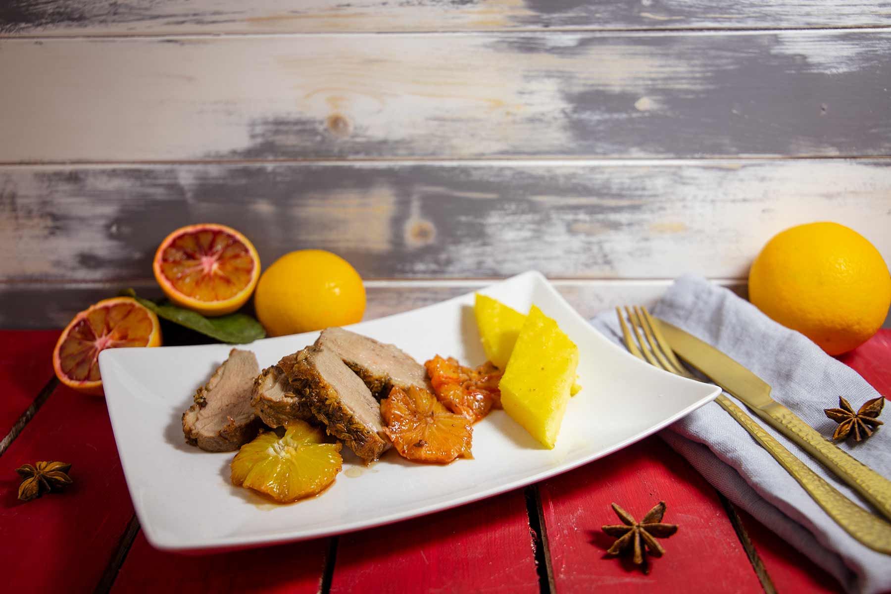 Vier Scheiben Entenbrust mit Blutorangenscheiben und zwei Polentastücken auf einem weißem Teller, daneben goldene Besteckgarnitur und ganze Zitrusfrüchte auf rotem Holztisch.