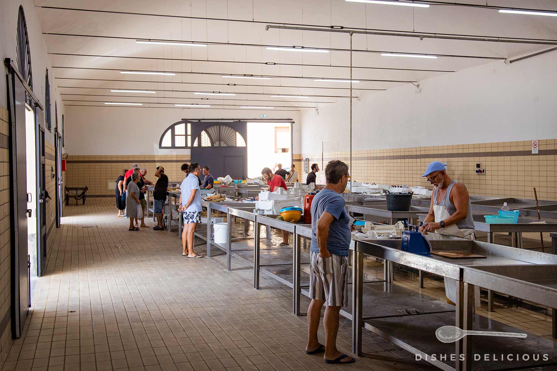 Fischmarkt in Trapani: Mehrere Personen stehen an langen Verkaufstischen in einem hellen, hohen Raum mit Kacheln an den Wänden und großen Fenstern.