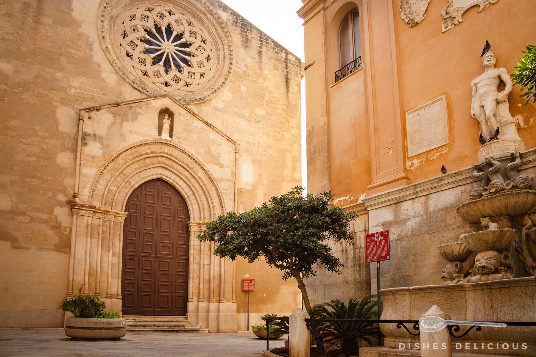 Kircheneingang mit rundem Rosettenfenster über einem großen, braunen Holztor, daneben ein Baum und der steinerne Saturn-Brunnen auf einem Platz in Trapani.