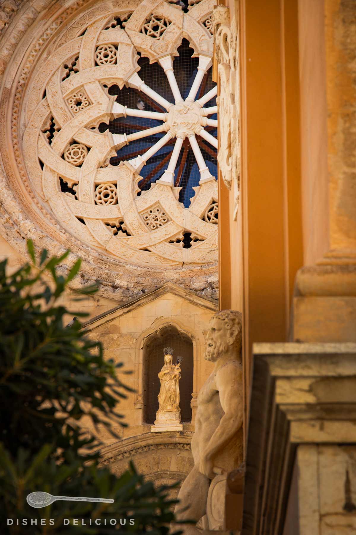 Rosettenfenster mit filigranen Steinornamenten und darunter eine Marienfigur in einer Nische, davor eine Saturn-Statue und Blätter eines Baums.