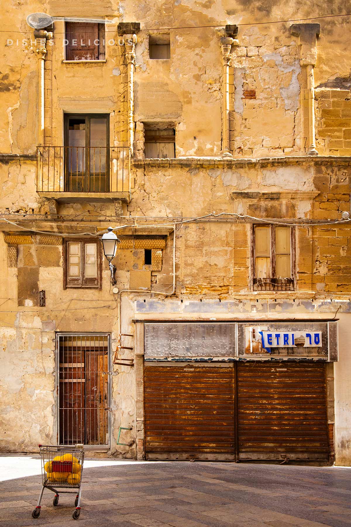 Verwitterte Fassade mit Balkon, mehreren Fenstern und zwei geschlossenen Holzläden, davor leerer Einkaufswagen mit gelbem Korb.