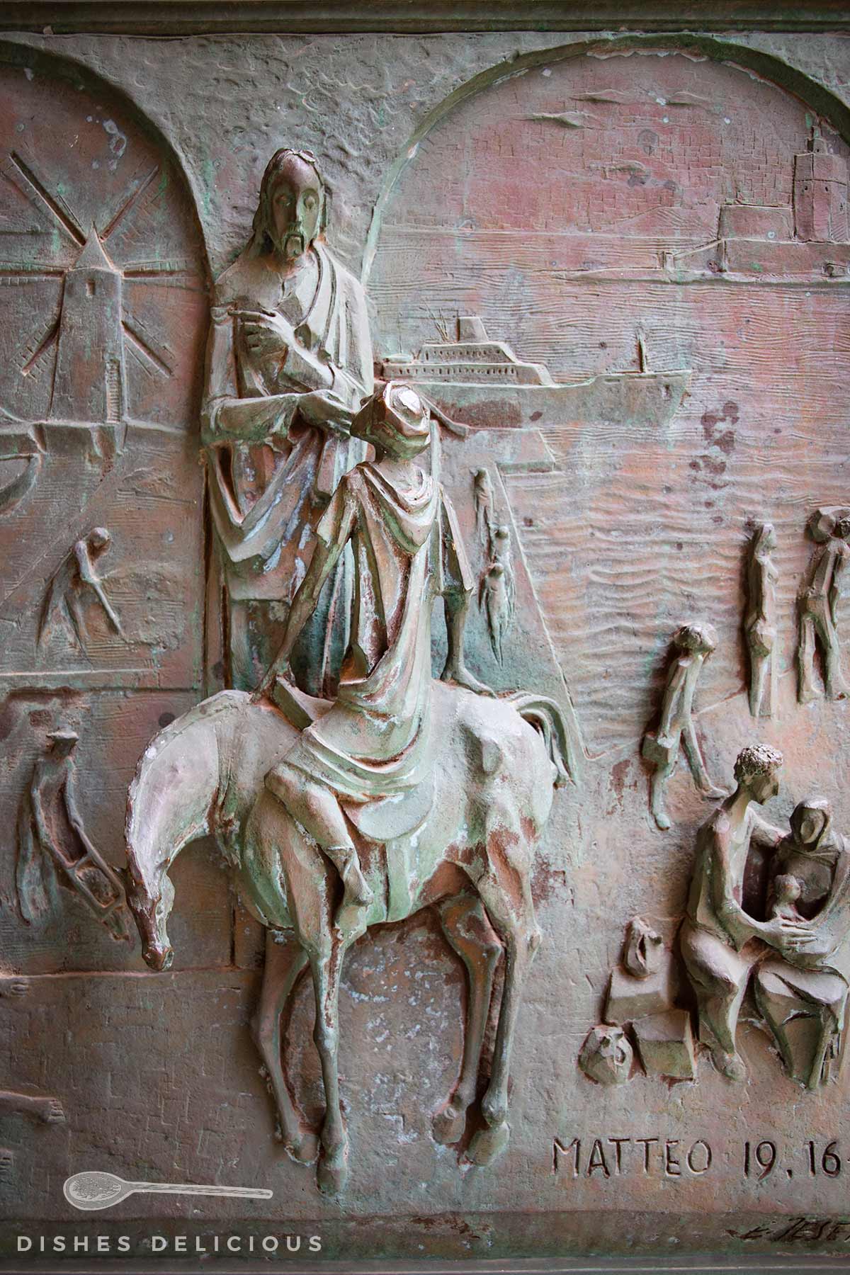 Nahaufnahme von Kirchenportal: Relief mit einer stehenden Figur, die eine sitzende Person auf einem Pferd hält, umgeben von weiteren Figuren und einem Schiff im Hintergrund, mit der Inschrift 'Matteo 19,16'.