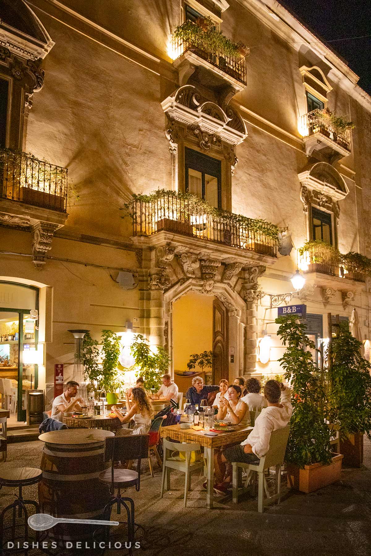Abendliche Straßenszene mit Gästen, die an Tischen vor einem historischen Gebäude mit Balkonen und Beleuchtung sitzen.