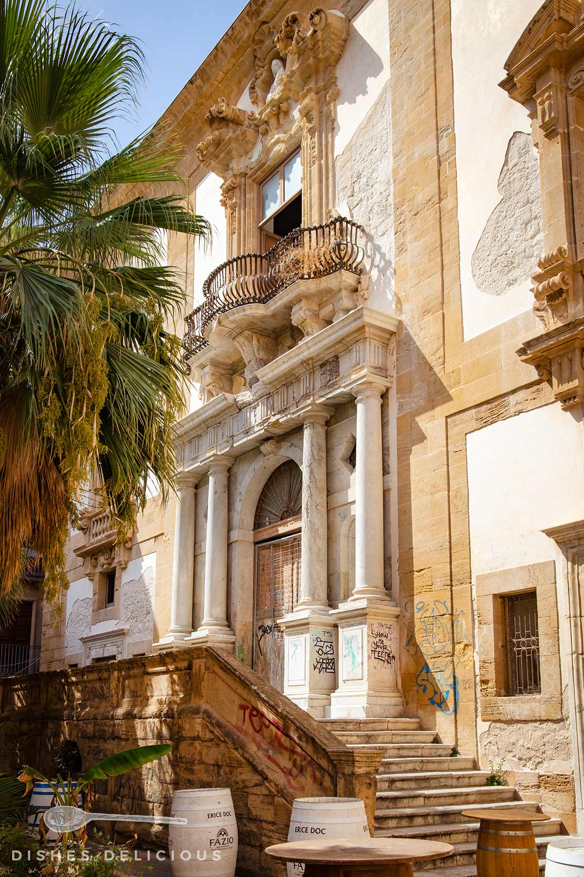 Barocke Sandsteinfassade mit Säulenportal, geschwungener Balkon und Treppe, umgeben von Palmen und Sitzgelegenheiten mit Graffiti an der Wand.