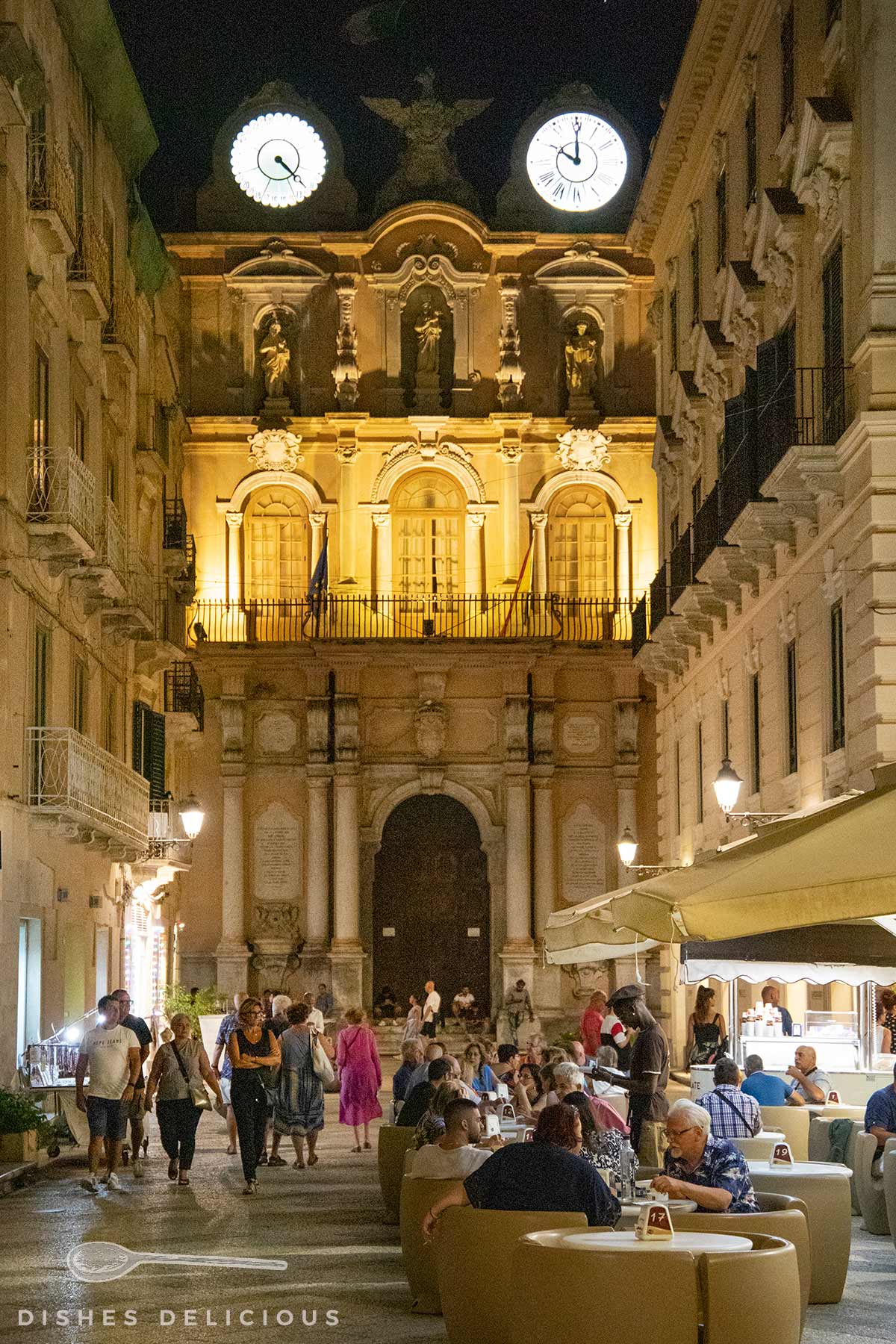 Belebte Straßenszene mit Sitzplätzen und Menschen vor beleuchtetem Palazzo Senatorio mit zwei großen Uhren und Statuen in der Fassade.