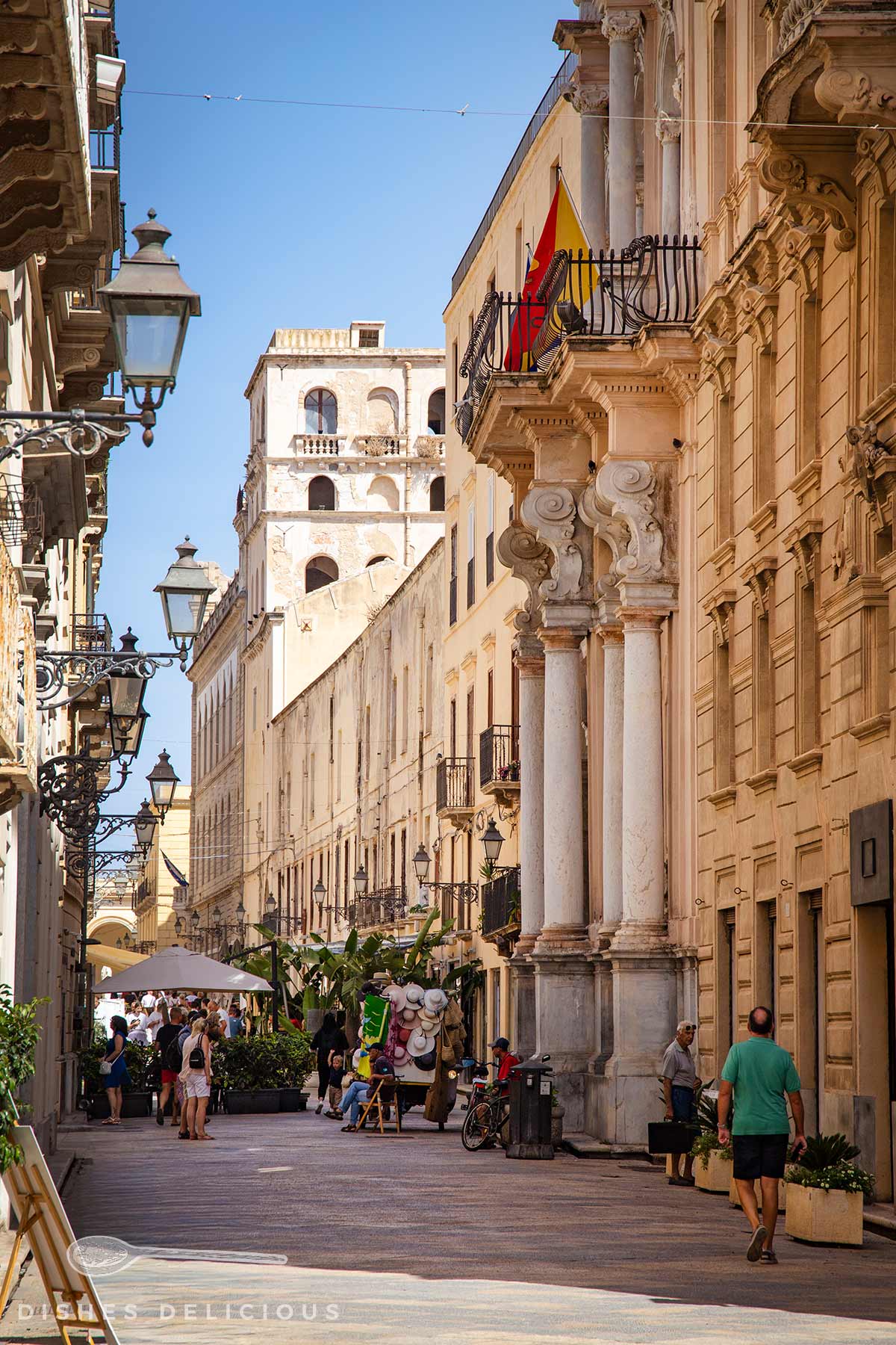 Altstadt-Straße mit historischen Gebäuden, Säulen und Balkonen, Menschen gehen und sitzen an Straßencafés, Sonnenschirme sichtbar.