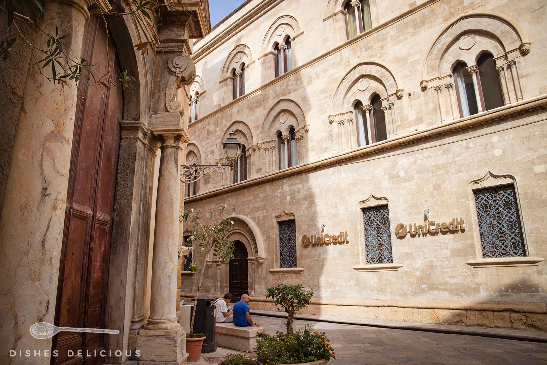 Historisches Gebäude mit UniCredit-Schriftzügen an der Fassade, zwei Personen sitzen auf einer Bank im Innenhof mit Pflanzen.