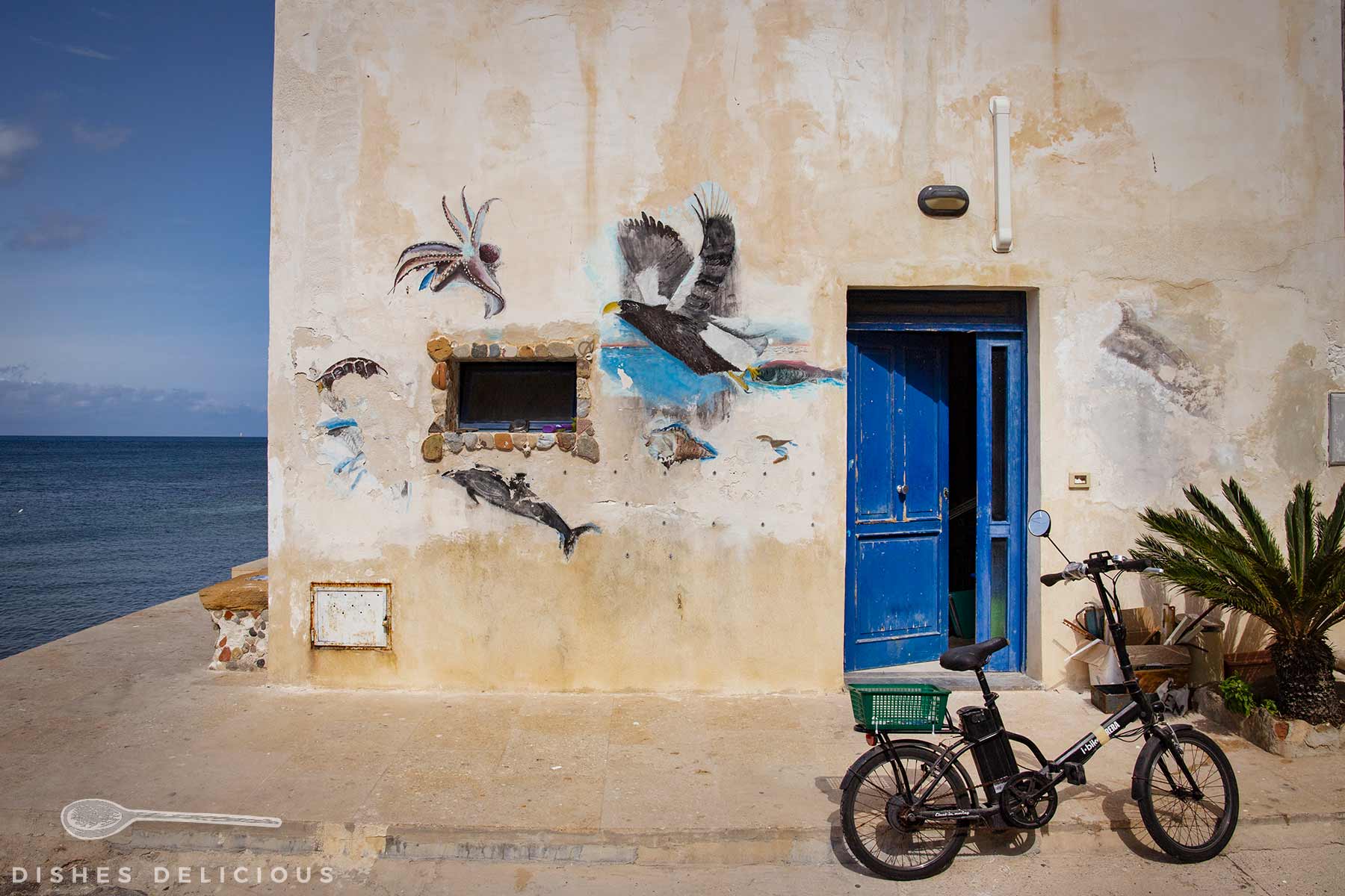 Fahrrad vor verwitterter Hauswand mit blauer Tür und Meeresmotiven als Wandmalerei neben Meerblick.