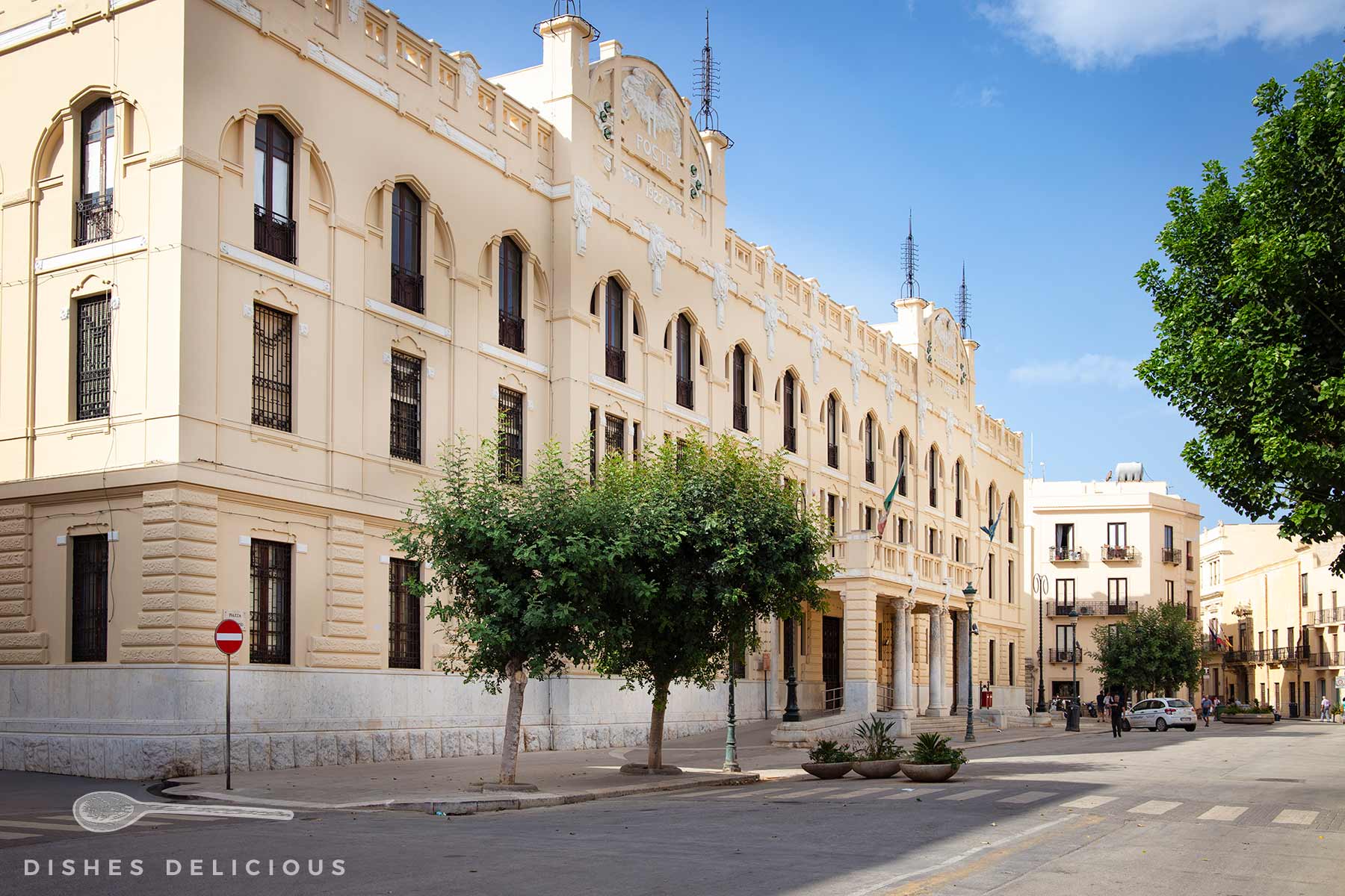 Historisches hellbeiges Postgebäude mit mehreren Fenstern und Bäumen davor an sonnigem Tag in einer ruhigen Straße.