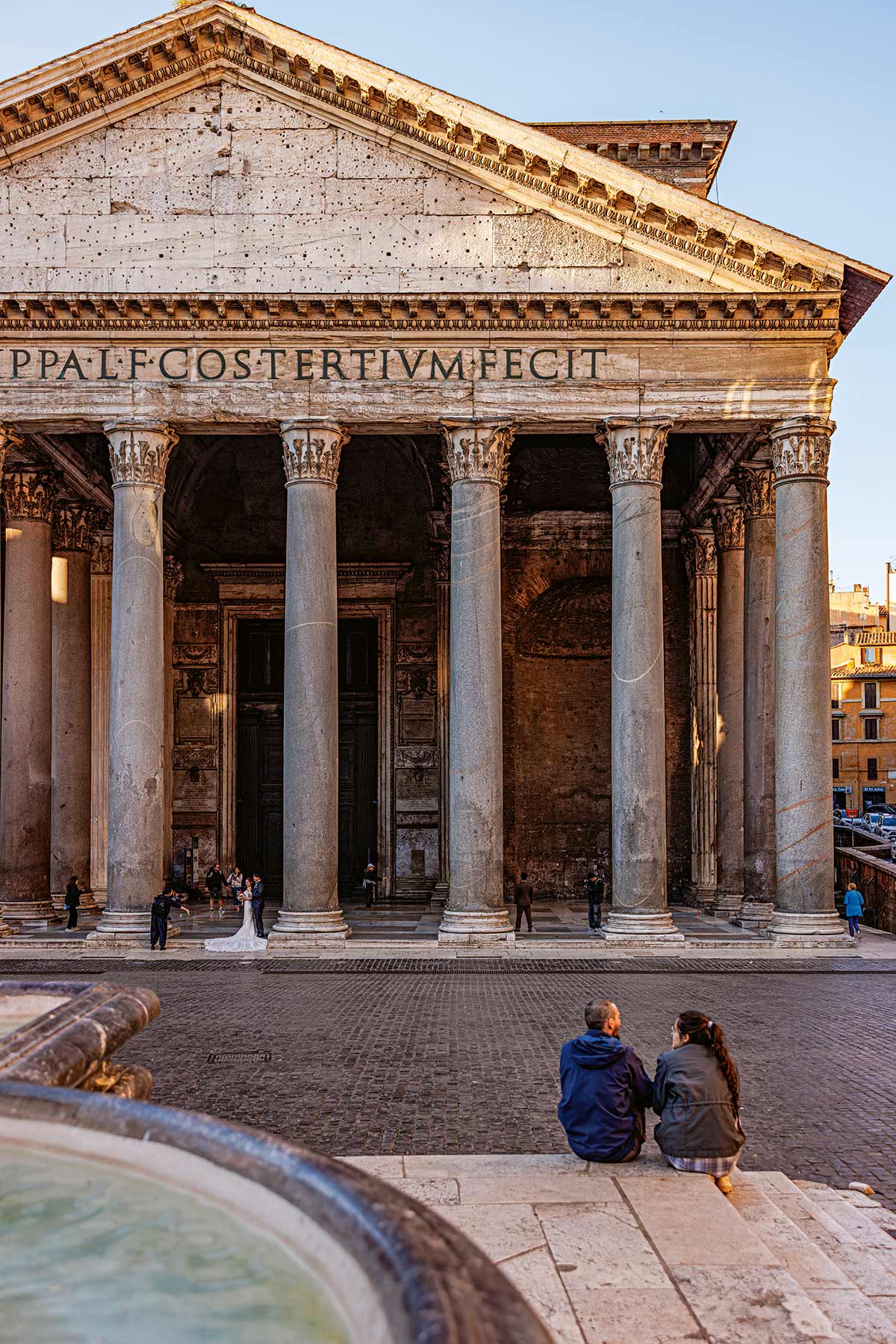 Vorderansicht des Pantheon in Rom mit sechs korinthischen Säulen und Inschrift im Giebel, zwei Personen sitzen im Vordergrund auf Stufen.
