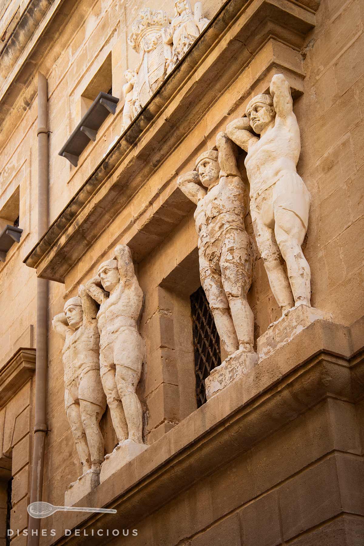 Vier steinerne Atlantenfiguren mit erhobenen Armen tragen einen Gebäudeteil an einer historischen Fassade aus Sandstein in Trapani.