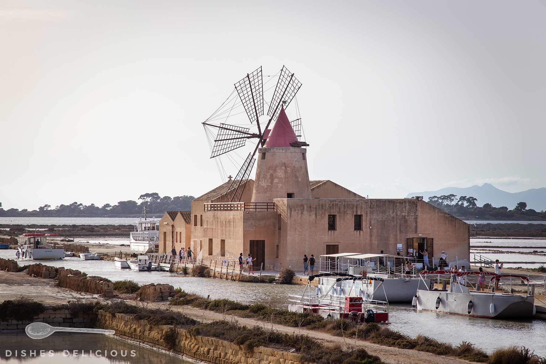 Windmühle der Saline Ettore e Infersa bei Trapani mit rotem Kegelhelm am Wasser, davor mehrere weiße Boote mit roten Sonnensegeln am Steg.