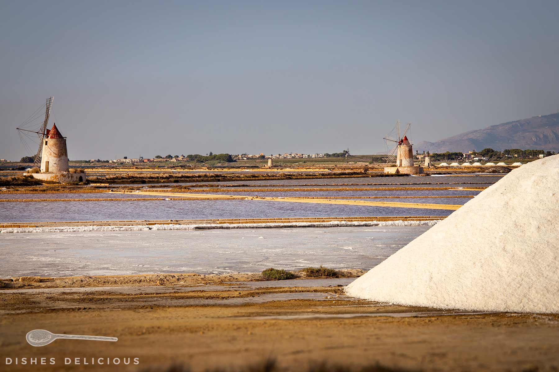Salzfelder mit zwei historischen Windmühlen und einem großen Salzberg im Vordergrund unter klarem Himmel.