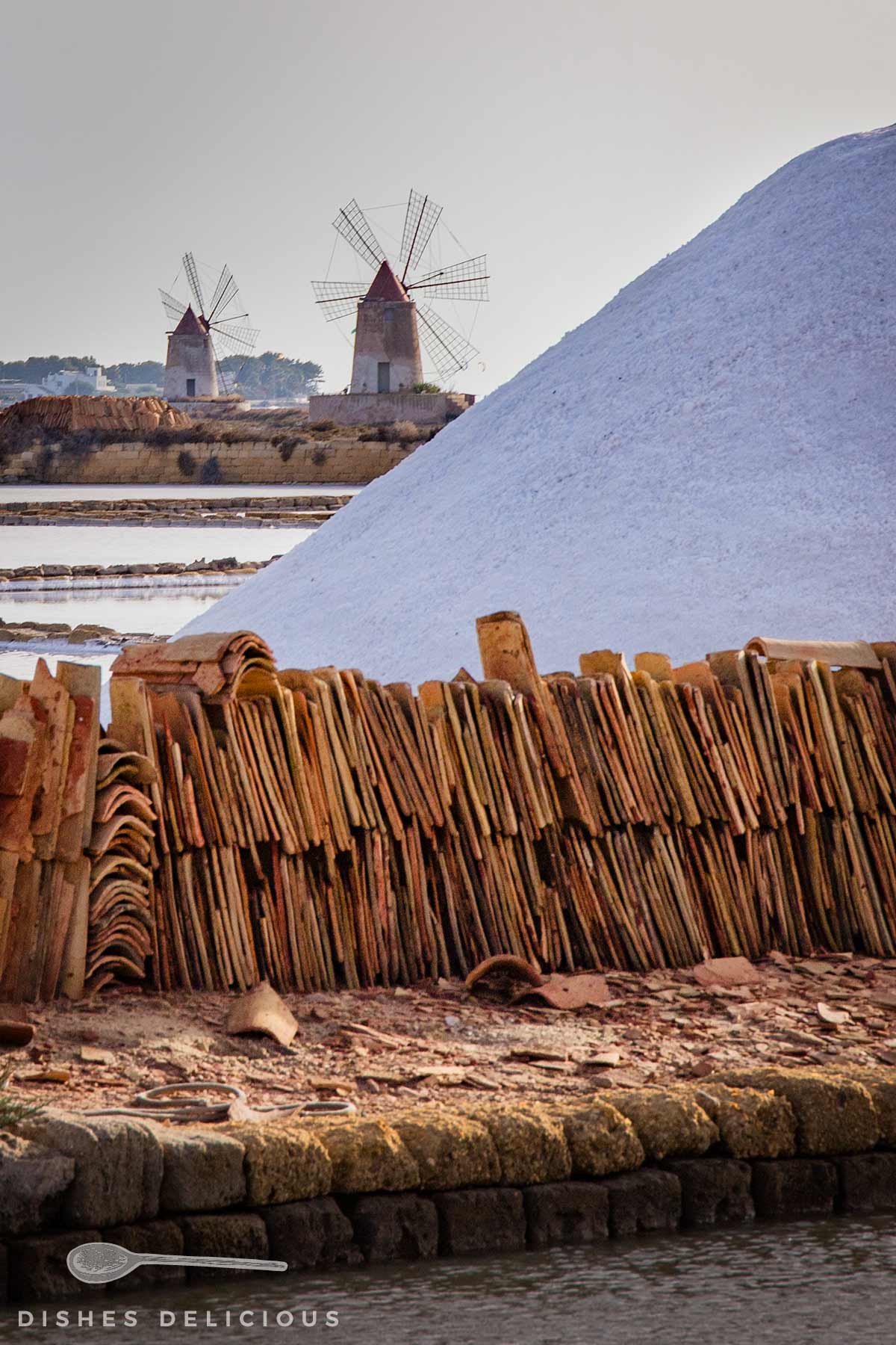 Großer Salzberg hinter einem Zaun aus gestapelten Ziegeln, im Hintergrund zwei historische sizilianische Windmühlen am Wasser.