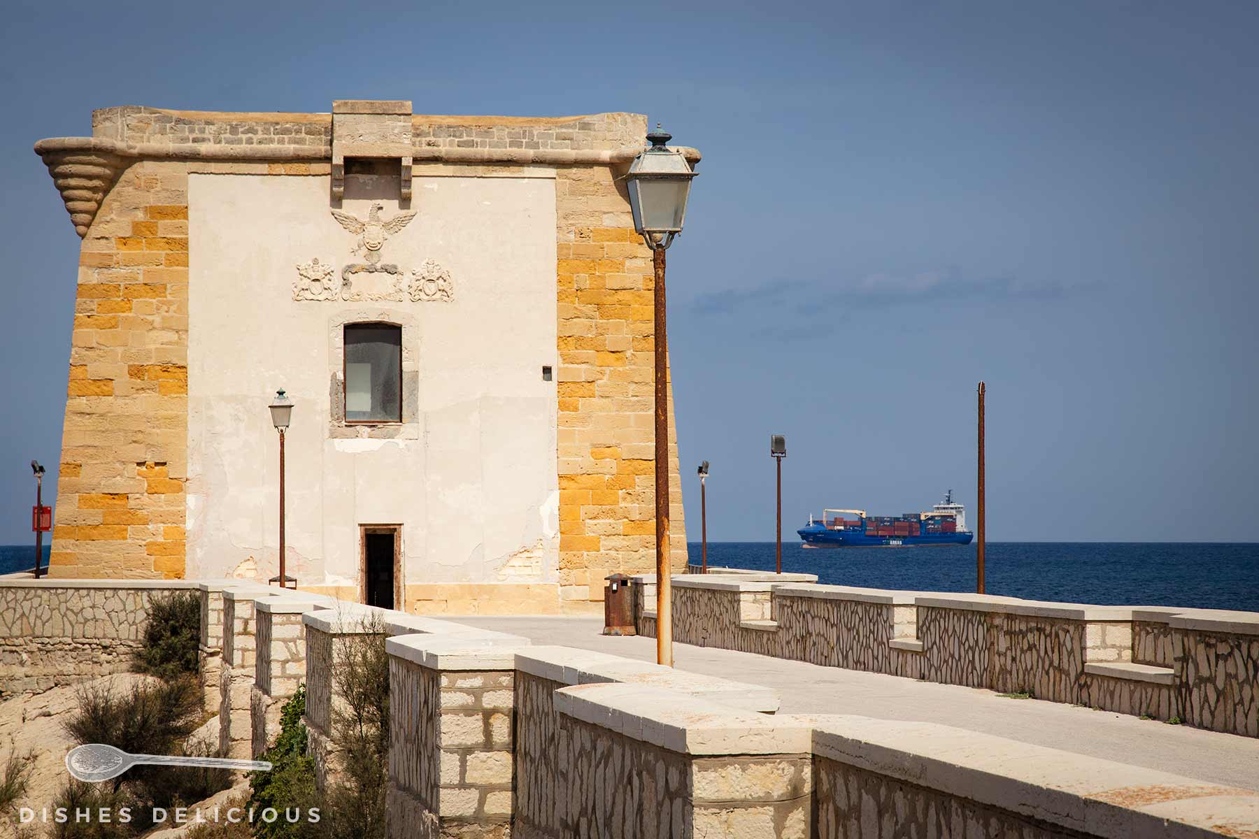 Alter steinerner Wachturm am Meer mit Laternen, dahinter ein Frachtschiff auf ruhiger See unter blauem Himmel.