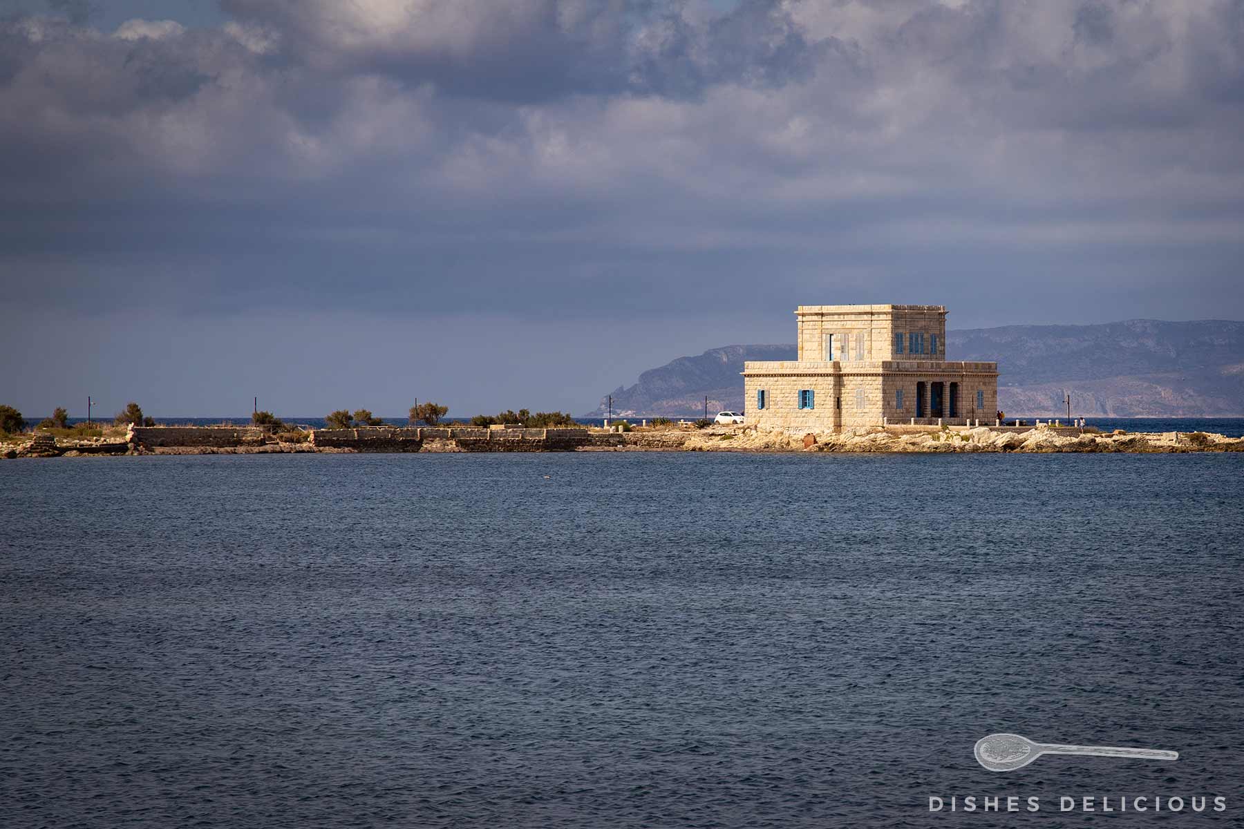 Rechteckiges, zweistöckiges Gebäude aus hellem Stein auf schmaler Landzunge im Meer unter bewölktem Himmel.