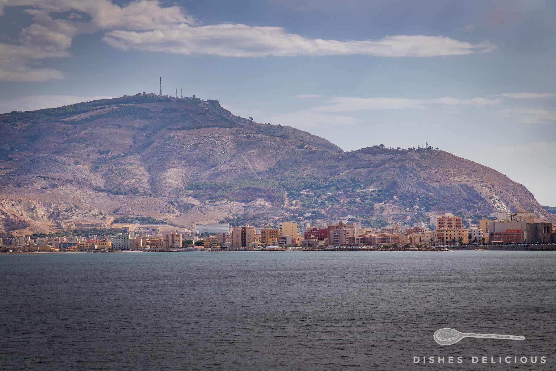Blick auf Trapani, Sizilien, mit mehrstöckigen Gebäuden. Im Hintergrund der bewaldete Berg Monte Erice unter bewölktem Himmel, im Vordergrund das Meer.