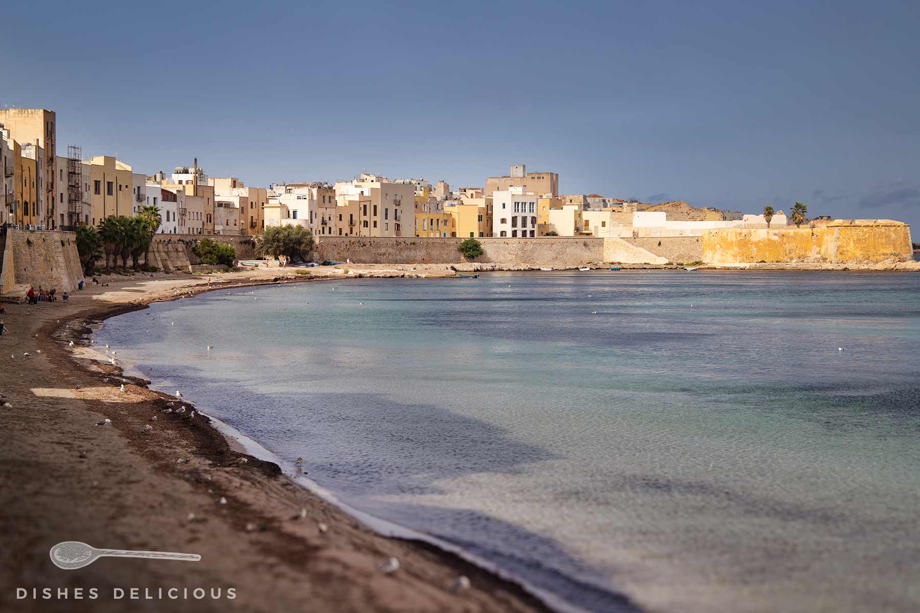 Altstadt von Trapani, Sizilien, mit Sandstrand und einer Stadtmauer, dahinter mehrstöckige, helle Gebäude unter bewölktem Himmel.