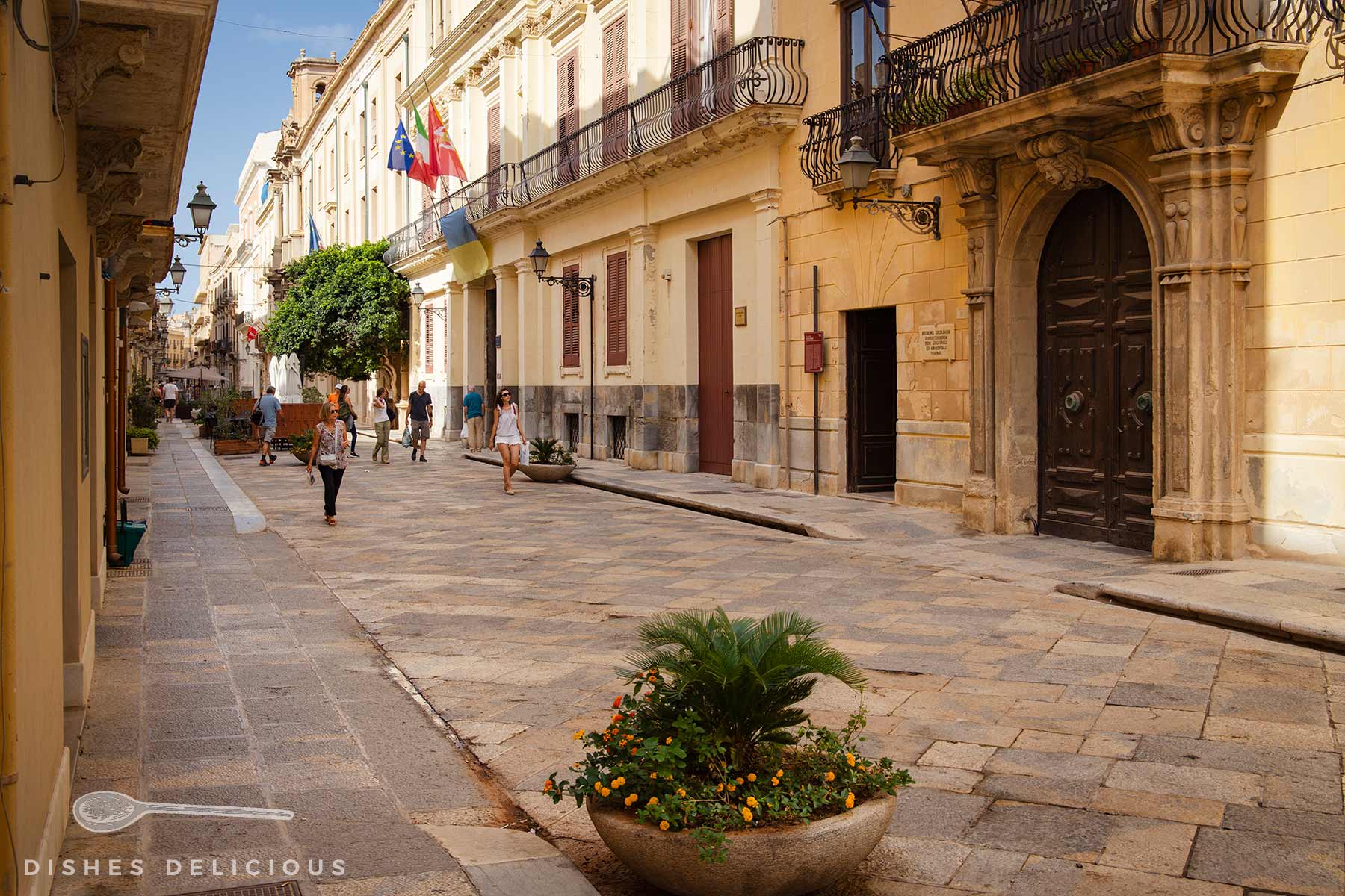 Via Garibaldi in Trapani mit historischen Gebäuden, mehreren Passanten und einem großen Pflanzgefäß mit grünen Pflanzen und gelben Blüten.