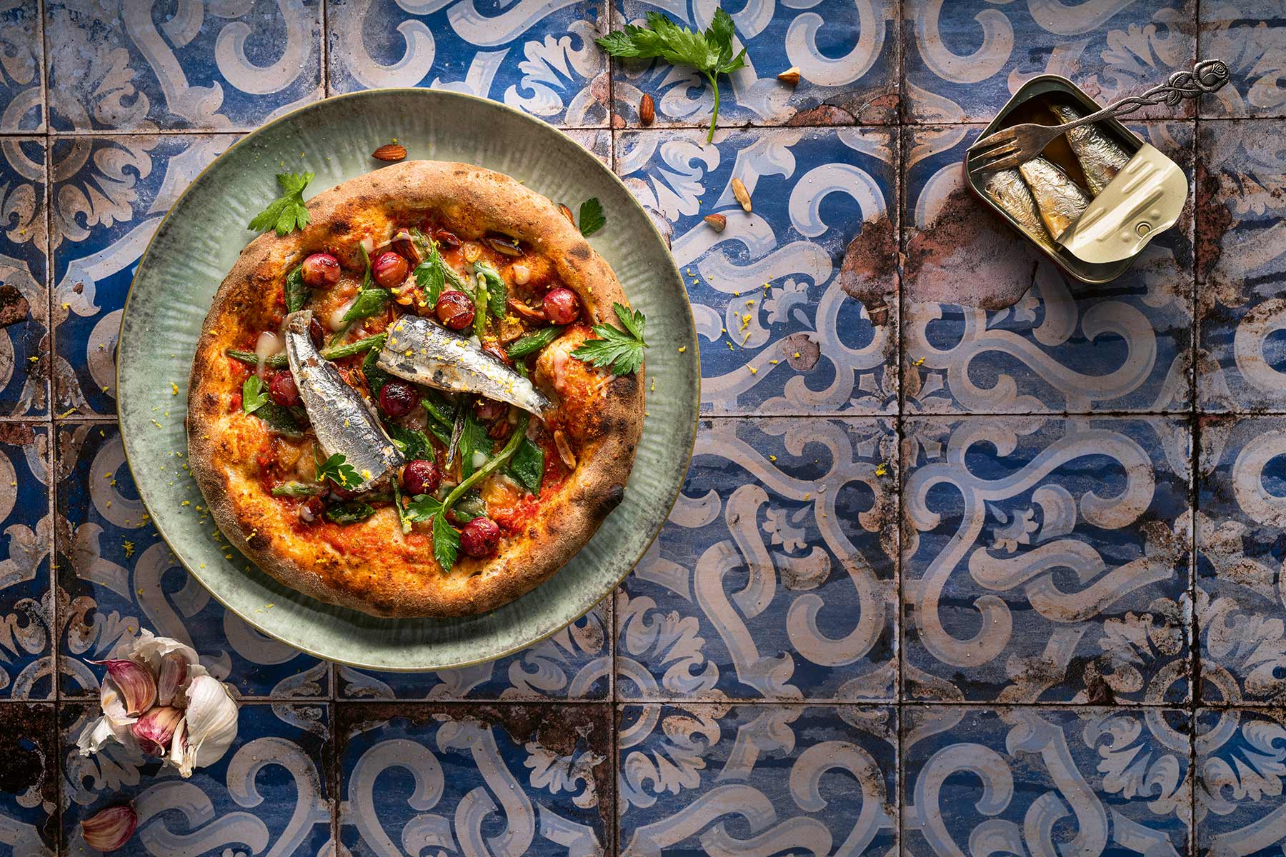 Pizza mit Sardinen, Kirschtomaten und Petersilie auf blau-weiß gemustertem Fliesenboden, daneben eine kleine Dose Sardinen und Knoblauchzehen.