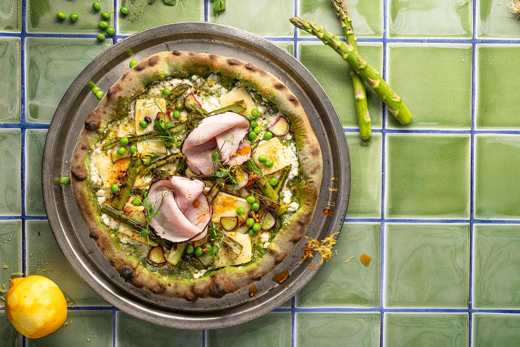 Pizza mit grünem Spargel, Erbsen, Schinken und Kräutern auf grün-blau gekacheltem Untergrund.
