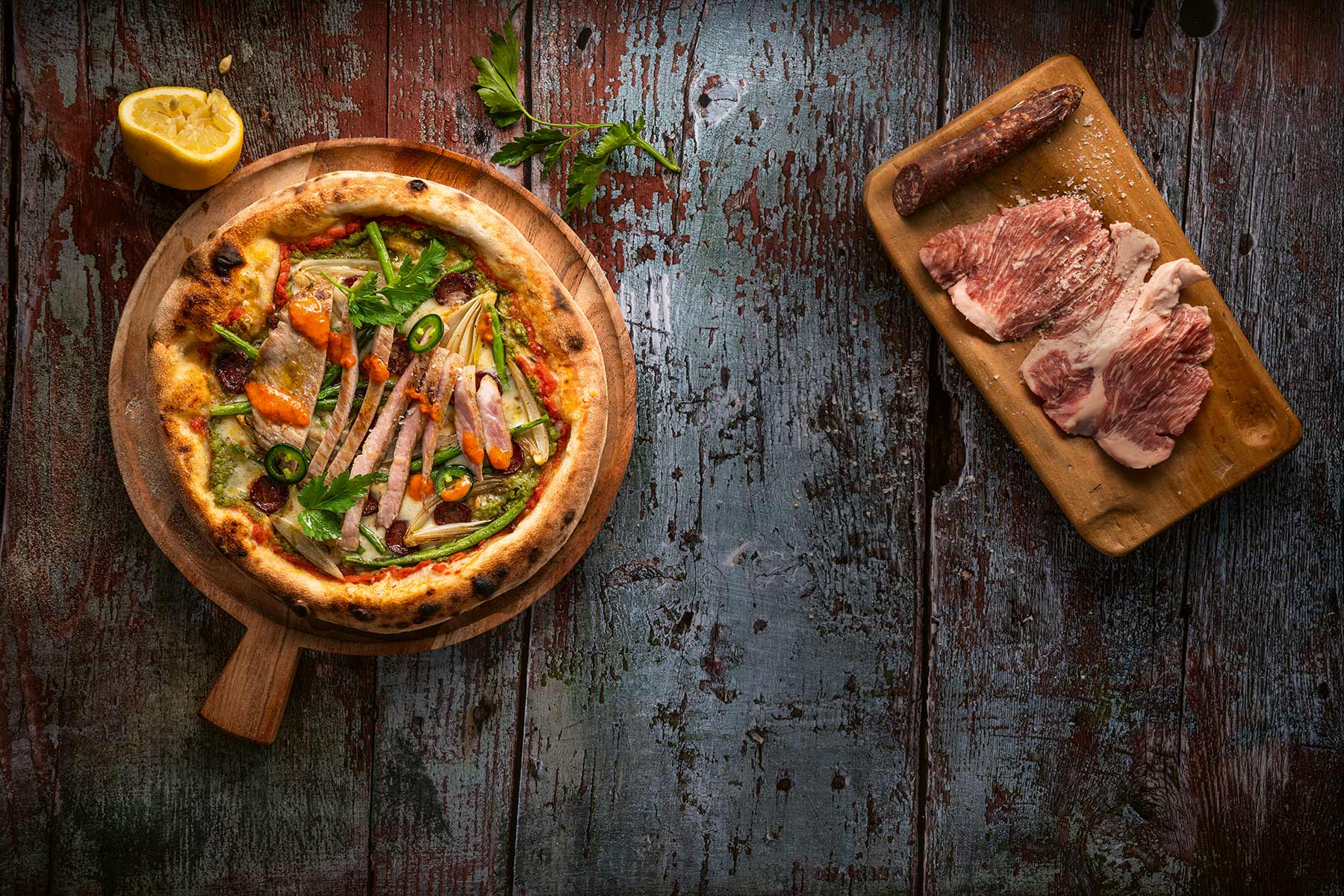 Pizza belegt mit feingeschnittenem Fleisch und grünem Gemüse auf einem Holzbrett. Daneben ein Brett mit geschnittenem Fleisch und einer Zitronenhälfte auf einem dunklen Holztisch.