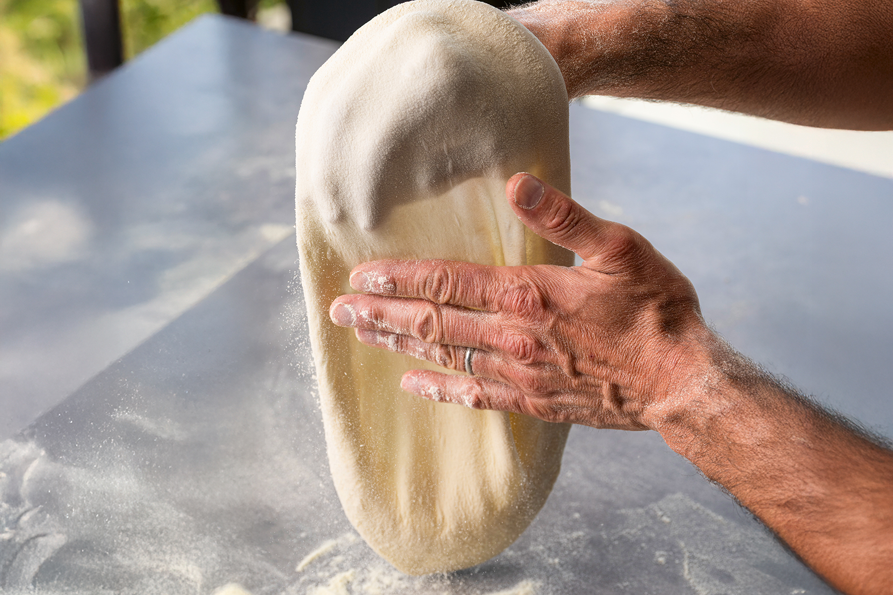 Hände formen einen dünnen, ovalen Pizzateig über einer bemehlten Arbeitsfläche.