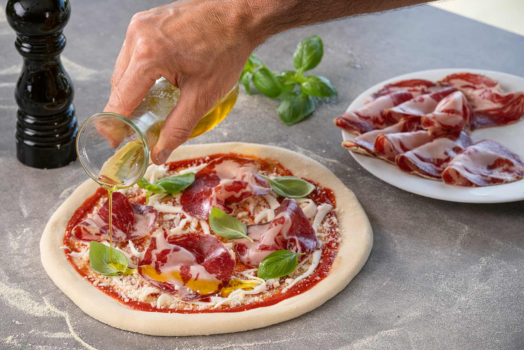 Jemand gießt Öl auf eine mit Tomatensauce, Käse, Basilikumblättern und rohem Schinken belegte Pizza.