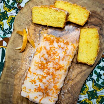 Sizilianischer Orangenkuchen mit Zuckerglasur und Orangenzesten, daneben drei geschnittene Kuchenstücke und Orangenschalen auf rustikaler Holzplatte.