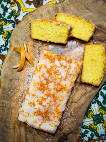 Sizilianischer Orangenkuchen mit Zuckerglasur und Orangenzesten, daneben drei geschnittene Kuchenstücke und Orangenschalen auf rustikaler Holzplatte.