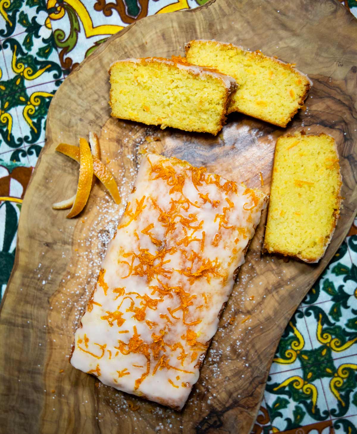 Sizilianischer Orangenkuchen mit Zuckerglasur und Orangenzesten, daneben drei geschnittene Kuchenstücke und Orangenschalen auf rustikaler Holzplatte.