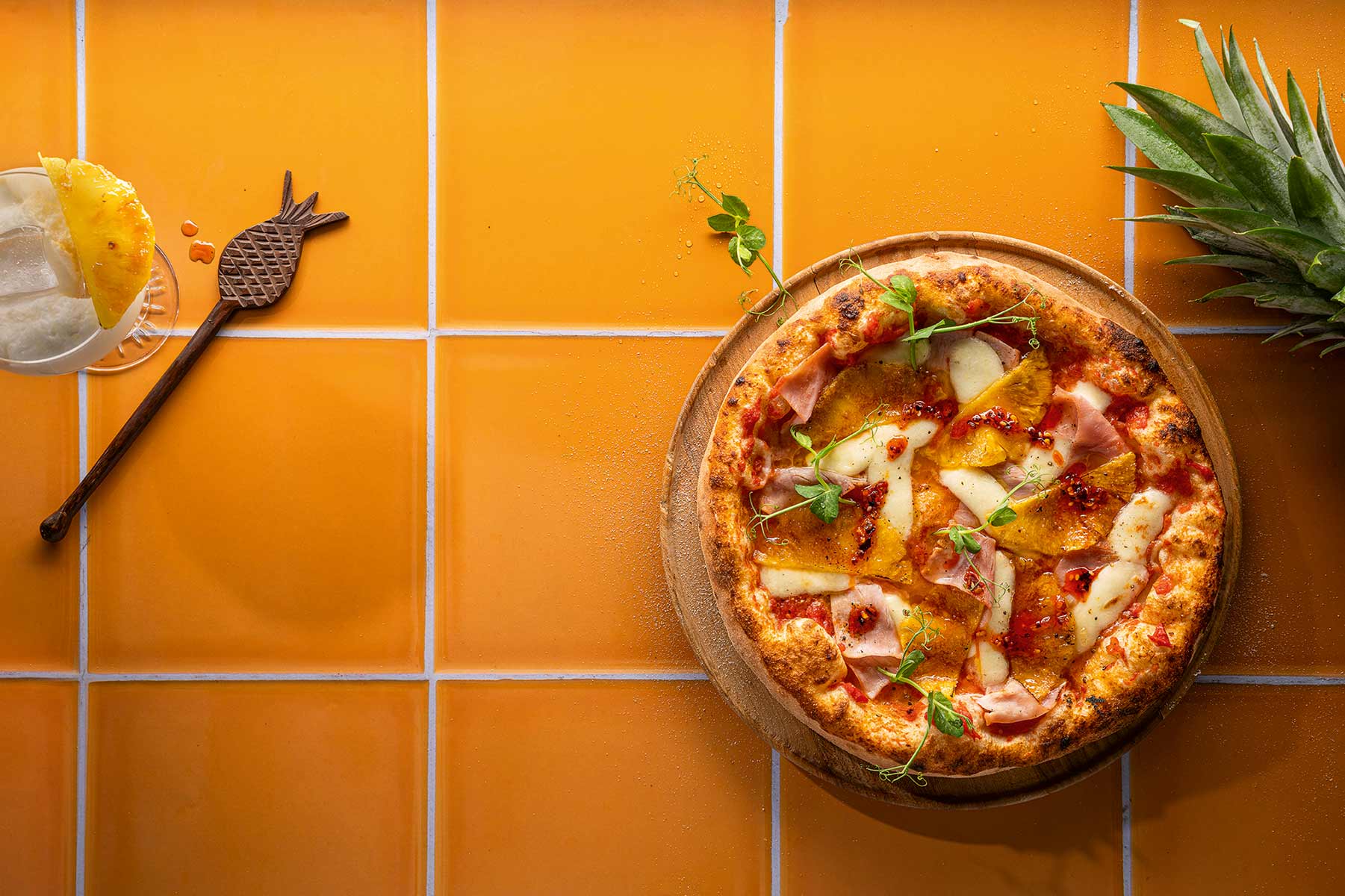 Pizza mit Schinken, Käse und Ananas auf rundem Holzbrett auf orangefarbenen Fliesen, daneben Ananasscheibe und Ananasblatt.