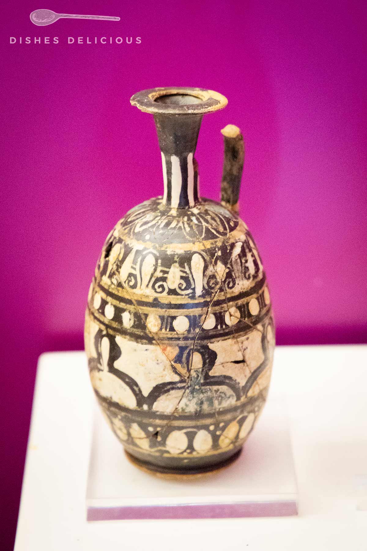 Antike Keramikkanne mit schwarzer Bemalung auf weißem Grund, schmaler Hals und Henkel, auf weißem Sockel vor violettem Hintergrund.