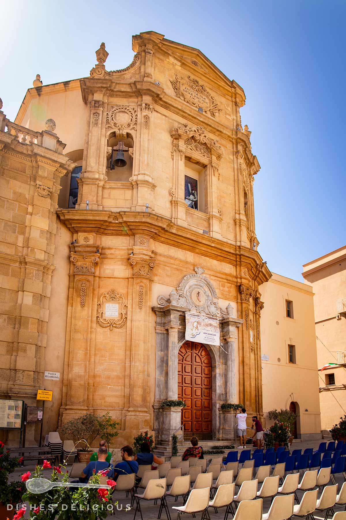 Barocke Kirchenfassade der Chiesa dell’Addolorata in Marsala mit verzierten Säulen, Rundbogenfenstern und Glocke, davor Stuhlreihen und Menschen im Freien.