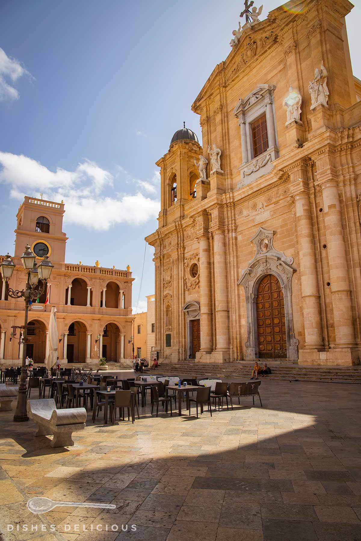 Sonniger Platz mit Tischen und Stühlen vor der barocken Mutterkirche von Marsala mit Skulpturen und großem Holztor.