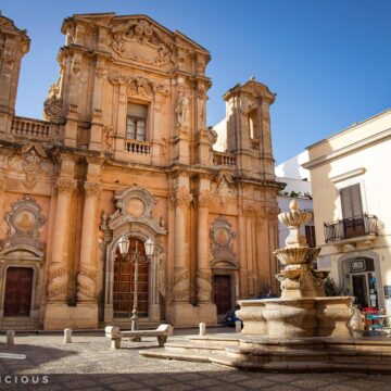 Barocke Fassade der Purgatorio-Kirche mit zwei Türmen und verzierten Säulen, davor ein steinerner Brunnen auf gepflastertem Platz mit Sitzbänken.