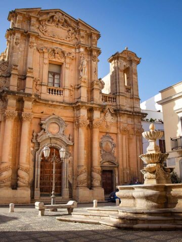 Barocke Fassade der Purgatorio-Kirche mit zwei Türmen und verzierten Säulen, davor ein steinerner Brunnen auf gepflastertem Platz mit Sitzbänken.
