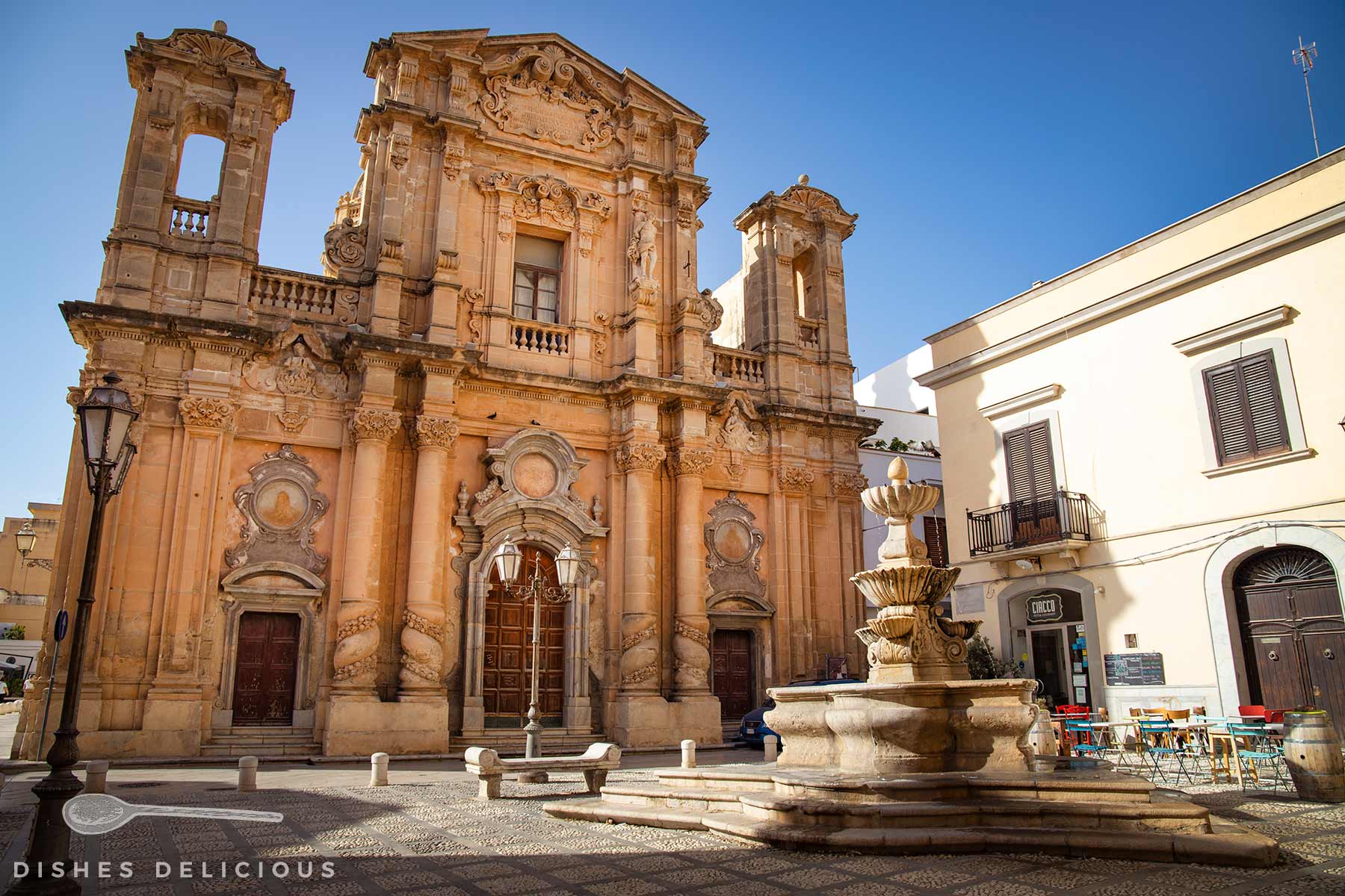 Barocke Fassade der Purgatorio-Kirche mit zwei Türmen und verzierten Säulen, davor ein steinerner Brunnen auf gepflastertem Platz mit Sitzbänken.